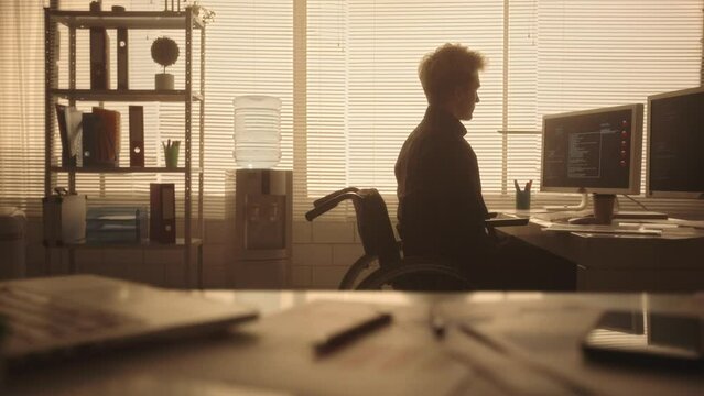 Silhouette of a young man in a wheelchair typing on a computer keyboard and drinking coffee in a dark office in the evening. A disabled manager works sitting at a table near a backlit window.
