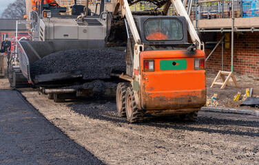 Asphalt paver filled with hot tarmac laying new road surface on new residential housing development site and roadworker operator in orange hi-viz next to it