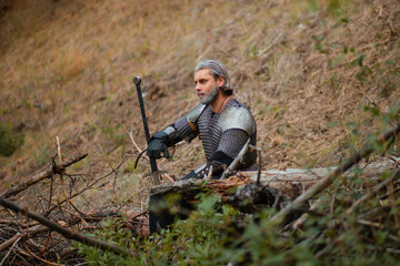 A gray-haired male warrior with a beard and a scar. In armor and with a sword, he sits on a log. An old knight at a halt with a weapon in his hands.