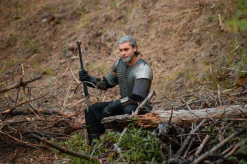 A gray-haired male warrior with a beard and a scar. In armor and with a sword, he sits on a log. An old knight at a halt with a weapon in his hands.