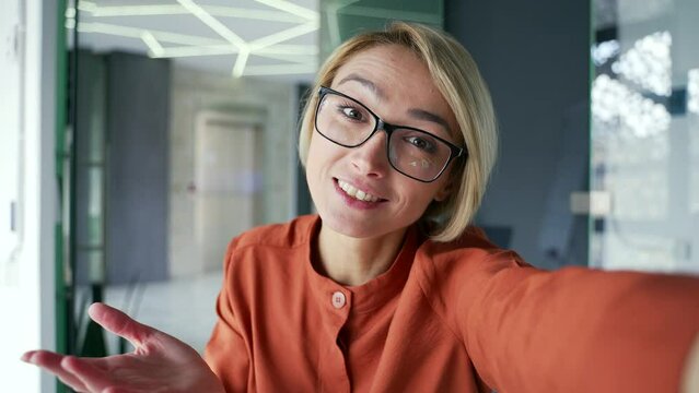 Webcam view. Yung smiling woman is having a video call using a smartphone while sitting at a workplace in a modern office. Happy female employee looking at camera, waving, laughing, talking to friend