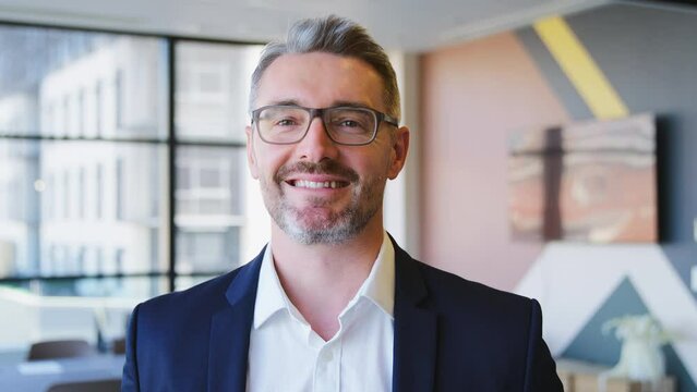 Portrait of smiling mature businessman wearing glasses standing in empty modern open plan office looking into camera - shot in slow motion