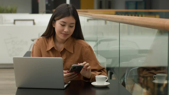 Asian woman businesswoman in cafe with laptop working with mobile phone scrolling social media smartphone chat online app chinese korean japanese freelancer student smiling girl studying in cafeteria