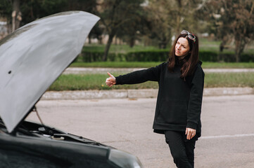 A young girl stands near a broken black car on a highway, road and asks for help from those passing by, raising her finger up, hitchhiking. Photography, portrait, breakage concept.