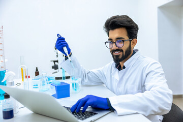 Scientist holds glass-dropper with reagents makes notes in laptop, experimenting with medical sample
