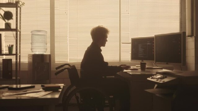 Silhouette of a young man in a wheelchair typing on a computer keyboard and drinking coffee in a dark office in the evening. A disabled male manager works sitting at a table near a backlit window.