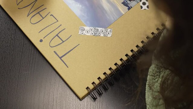 Video of an anonymous hand drawing with a marker pen the word Thailand in Spanish on the cover of a papercraft album.