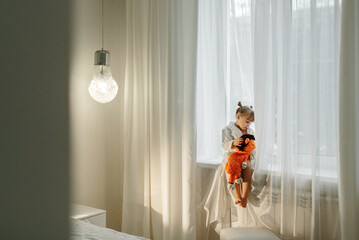 A girl in a bathrobe plays on the windowsill with a knitted lion