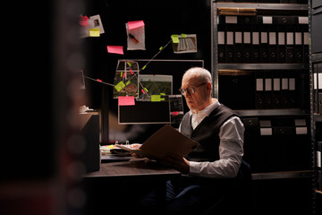 Senior police officer looking at criminal investigations files, working overhours at mysterious suspect profile in arhive room. Elderly private detective analyzing crime scene evidence