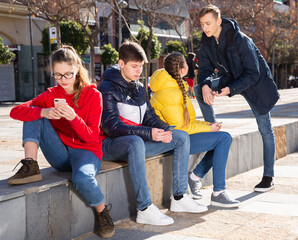 Four teenagers having friendly discussion and using cellphones during gathering outdoors on spring day