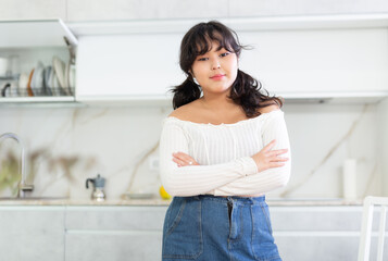 Fototapeta na wymiar Portrait of smiling young housewife in white blouse and denim skirt standing in kitchen, posing on camera