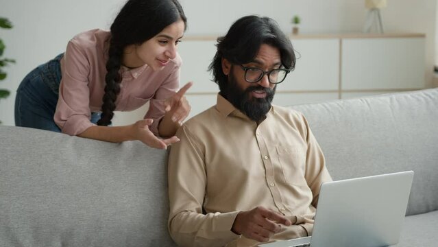Arabian muslim man use laptop app Indian woman behind husband talk excited clap hands shopping online happy diverse couple choose goods make internet order search apartment rent home on estate website