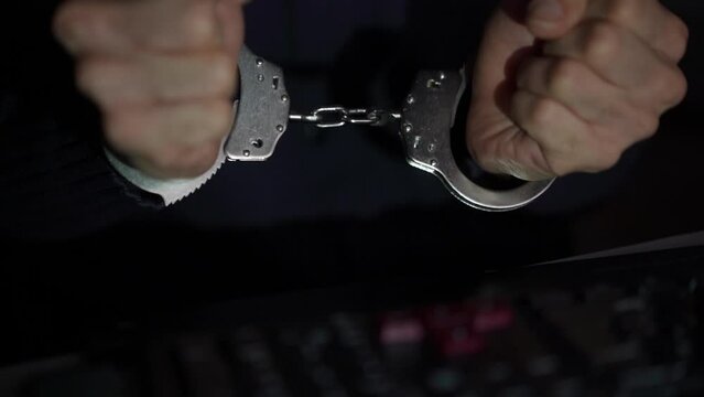 Handcuffed hacker arrested by police. Police siren in the background.Hands tied with chains on wrists typing on laptop keyboard. 