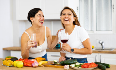 Obraz na płótnie Canvas Two happy female friends preparing salad and drinking red wine together in modern kitchen