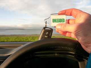 Obraz premium Driver holding car key with unleaded badge. Beautiful nature scene with ocean and mountain out of focus in the background. Travel or trip in rented car concept.