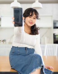 Smiling young girl showing modern smart phone with blank screen, in the living room or in the kitchen