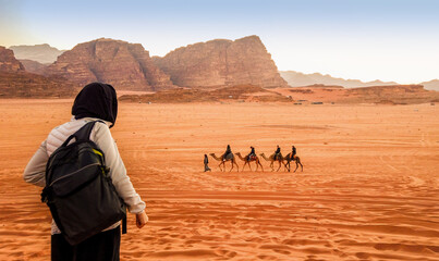 Jordan, Wadi Rum. Caravan of camels with drovers in the desert, rock mountains at sunset. Tourist from behind looks at the panorama. - obrazy, fototapety, plakaty