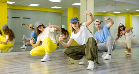 Group portrait of talented active tween dancers in casual clothes squatting and learning new dance move in hall