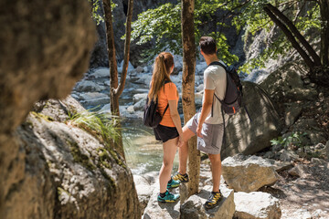 Two tourists, a young couple, enjoying time in nature and hiking, walking along the rocky mountain stream bed and looking in the distance