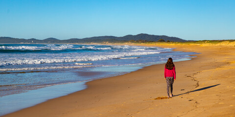 beautiful long haired girl walking on wooli beach during sunrise; long beach with large waves on the coast of pacific in yuraygir national park, new south wales, australia