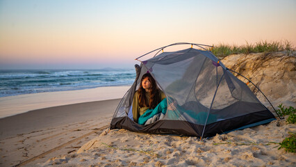 A beautiful girl admires a wonderful sunrise on the beach from transparent tent. Camping on the beach in Australia, Hat Head National Park, NSW