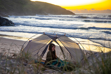 A beautiful girl admires a wonderful sunrise on the beach from her transparent tent. Camping on the beach in Australia, Hat Head National Park, NSW