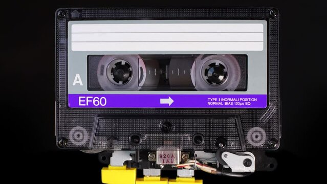 A Purple, vintage, compact cassette tape playing back in a player mechanism. Tape recorder head and rotating reels of deck player. Magnetic tape stores stereo music in a magnetic image
