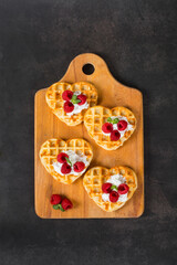Viennese heart-shaped waffles with cream and raspberries. On a serving wooden board. Dark gray background. Top view