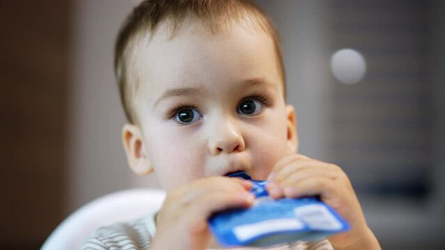 Lovely Caucasian baby boy eating the fruit puree. Beautiful kid holding doy pack in both hands sucking the food. Close up.