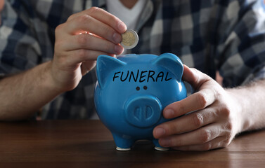 Money for funeral expenses. Man putting coin into piggy bank at wooden table, closeup