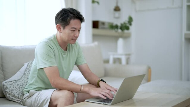 Young Asian man working on laptop computer while sitting at the living room.