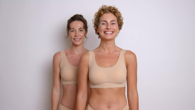 Two attractive seminude models wearing ergonomic beige colour bras standing on white background, both smiling at camera, comfortable underclothing concept, copy space