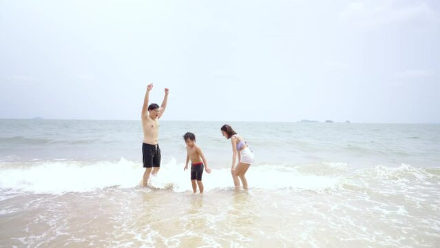 Happy Asian family travel ocean on summer holiday vacation. Parents and little son in swimwear have fun outdoor activity lifestyle walking and playing sea water together at tropical beach in sunny day