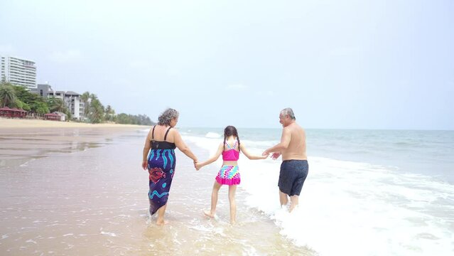 4K Happy Asian family travel ocean on summer holiday vacation. Grandparents and grandchildren girl in swimwear enjoy and fun outdoor lifestyle playing sea water together at tropical beach in sunny day