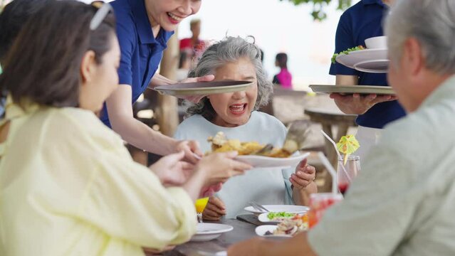 Happy Asian couple and aging parents having lunch together during travel tropical beach on summer holiday vacation. Waiter and waitress serving food and drink to tourist customers at beach restaurant.