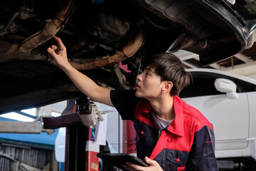 Fototapeta na wymiar Professional young Asian male motor mechanic inspects undercarriage of electric car(EV) lifted by forklift jack for repair at garage, automotive maintenance service works industry occupation business.