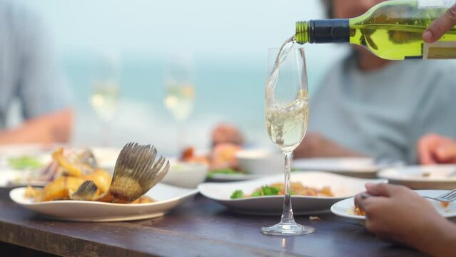 Happy Asian family couple and aging parents having celebration dinner during travel ocean together on summer holiday vacation. Waiter and waitress serving champagne to customers at beach restaurant.