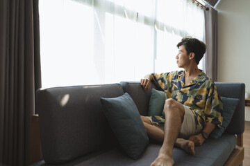 Tourist young adult Asian man smile, feel relaxed, happy, sitting on hotel sofa when look at beautiful scene on window at hotel room. Traveler relax on sofa when travel on holiday trip. Travel concept - 594876535