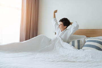Asian tourist man in bathrobe is waking up on bed, stretching on bed for fresh at bedroom during travel trip in morning in hotel bedroom. Tourist Asian guy get up in morning. Waking and get up concept - 594876544