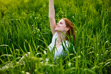 a cute woman in the summer high grass sits in a light dress happily raising her hand up