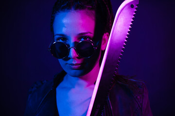 Young woman in a leather jacket in the dark with a machete knife in her hands. Neon light blue pink...