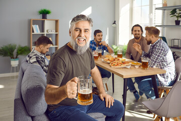 Portrait of attractive mature bearded man holding a glass of beer looking at camera and smiling with a company of cheerful happy male friends talking in the background. Men enjoying party together.