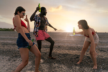 Beach Party with Friends at Sunset - Two Caucasian women, African man in twenties, drinking beer, dancing, backlit shot, swimwear and summer clothes.