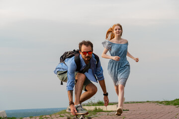 Positive young man riding on longboard while his girlfriend running near him against gray sky