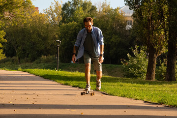 Fototapeta premium Front view of young guy riding on longboard along empty asphalt road between green trees