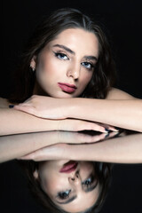 Studio portrait of beautiful brunette woman with long wavy hair and nude makeup. Model placed head on hands. Mirror reflection of model on glass surface. Toned image with blue color