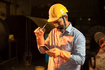 Portrait of happy Indian male Mechanical Engineer wearing yellow safety helmet and gloves holding smart phone in hand in Industrial factory, Skill india.