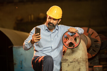 Portrait of happy Indian male Mechanical Engineer wearing yellow helmet holding smart phone in hand in Industrial factory, Skill india.