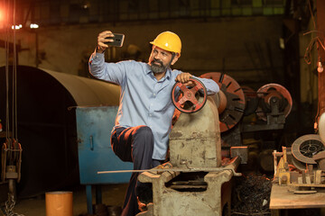Portrait of happy Indian male Mechanical Engineer wearing yellow helmet holding smart phone in hand...