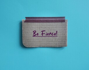 Card box paper stick on blue background with handwritten text BE FIERCE!, concept of affirmation to...